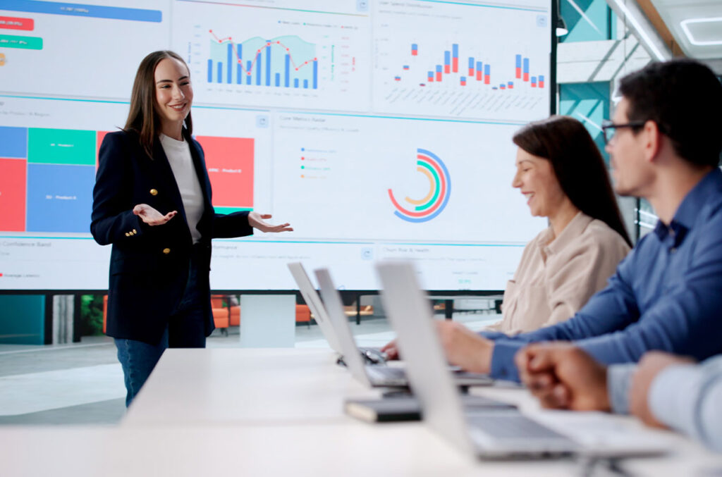 Person presents data dashboards on a large screen while colleagues review charts and laptops during a meeting.