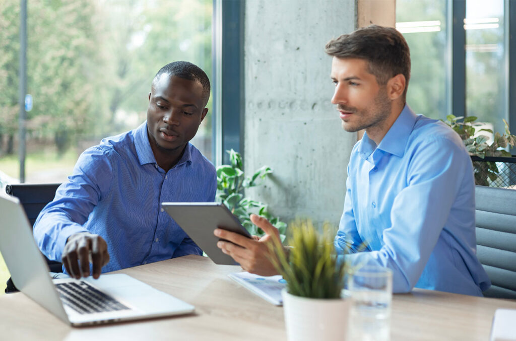 Two professionals review information on a laptop and tablet while meeting at a conference table in a modern office.