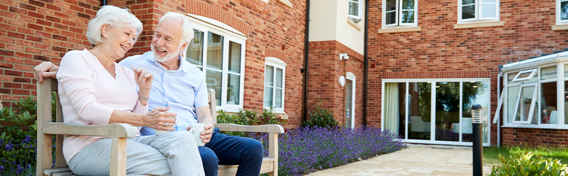 Two older adults relax and talk on a bench outside a senior living community.