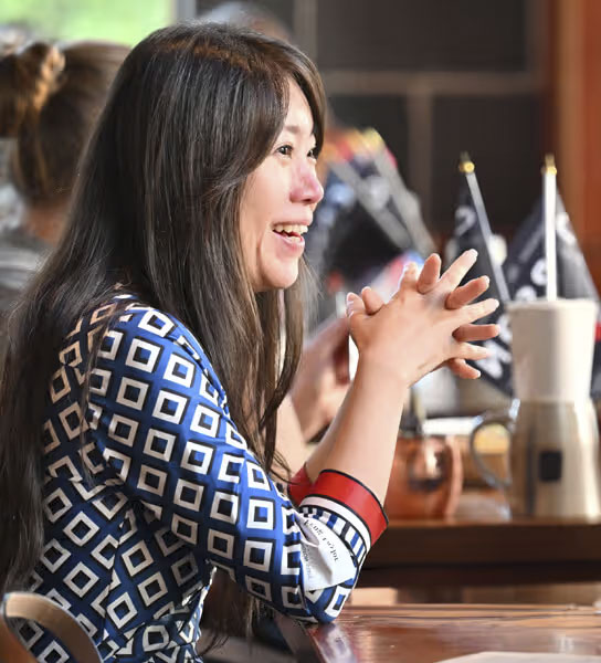 A person with long hair sitting at a table, wearing a blue and white geometric patterned shirt with red cuffs. In the background, there are other people and some small flags on the table.