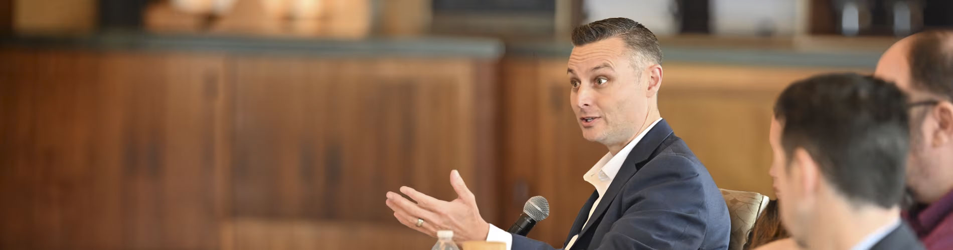 Three individuals seated at a table, engaged in a discussion. One person is speaking into a microphone and gesturing with their hand. The background features wooden paneling and blurred objects on shelves.