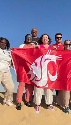 A group of people standing on sand in Dubai, holding a red flag with a white WSU Cougar head logo. The background is a clear blue sky.