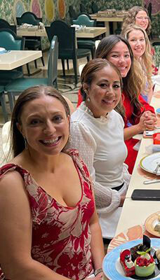 A group of people seated at a dining table for the welcome dinner, likely at a restaurant. The table is set with plates, cutlery, and various dishes. The background includes more tables and chairs, as well as decorative elements on the walls.