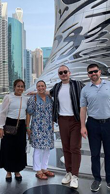 Four people standing on the view deck of the Museum of the Future in Dubai. The background features part of the museum's futuristic architecture with its distinctive silver, curved design and Arabic calligraphy. Several high-rise buildings are visible in the distance, including a prominent skyscraper with a unique triangular top.