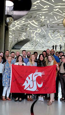 A group of people posing for a photo inside the Museum of the Future in Dubai. The group is holding a red flag with a white WSU Cougar head logo. The background features the museum's distinctive futuristic design with Arabic calligraphy and intricate patterns on the ceiling.