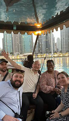 A group of people on a boat ride at the Dubai Fountains. The background features tall buildings and illuminated fountains, indicating it is evening or night time. The people in the boat are taking selfies and enjoying the ride.
