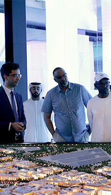 A group of people standing and observing a detailed architectural model of a city, representing Masdar City in Abu Dhabi, Dubai. The background includes large screens or windows with bright lighting.