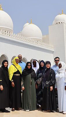 A group of people standing in front of the Sheikh Zayed Grand Mosque in Abu Dhabi, UAE. The mosque's distinctive white domes and intricate architectural details are visible in the background. The people are dressed in traditional attire, with some wearing abayas and others wearing thobes.