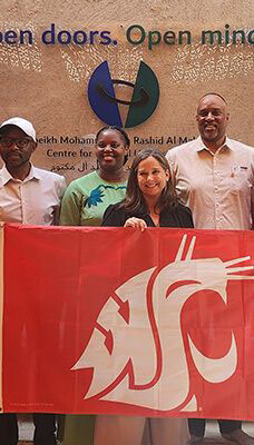 A group of people standing in front of a wall with the text "Open doors. Open minds." and "Sheikh Mohammed bin Rashid Al Maktoum Centre for Cultural Understanding" written on it. The people are holding a red flag with a white WSU Cougar head logo.