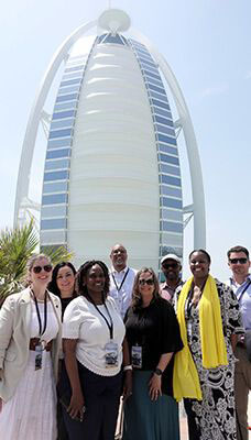 A group of people standing in front of the Burj Al Arab in Dubai. The Burj Al Arab is a luxury hotel known for its distinctive sail-shaped silhouette. The people in the image are posing for a photo with the iconic building prominently visible behind them.