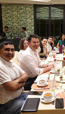 A group of people seated at a dining table for the welcome dinner, likely at a restaurant. The table is set with plates, cutlery, and various dishes. The background includes more tables and chairs, as well as decorative elements on the walls.