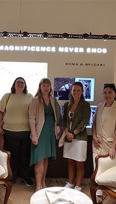 Four people standing inside a Bulgari store in Italy. The background features a lit sign with the text "MAGNIFICENCE NEVER ENDS" and "ROMA o BVLGARI." There are also two display screens showing images of jewelry or watches. The people are standing in front of the displays, and there are chairs visible in the foreground.