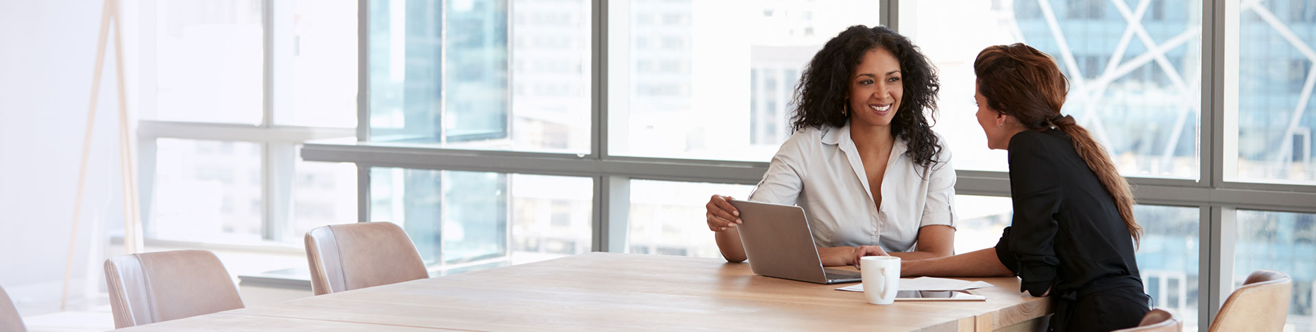 Two people sitting at a long wooden table in a modern office setting with large windows and city buildings visible outside.