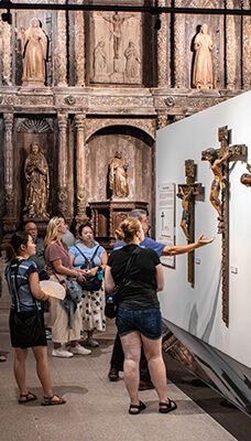 A group of people inside the Casa Manila Museum, observing religious artifacts, including crucifixes displayed on a white wall. The background features an ornate wooden altar with statues and carvings.