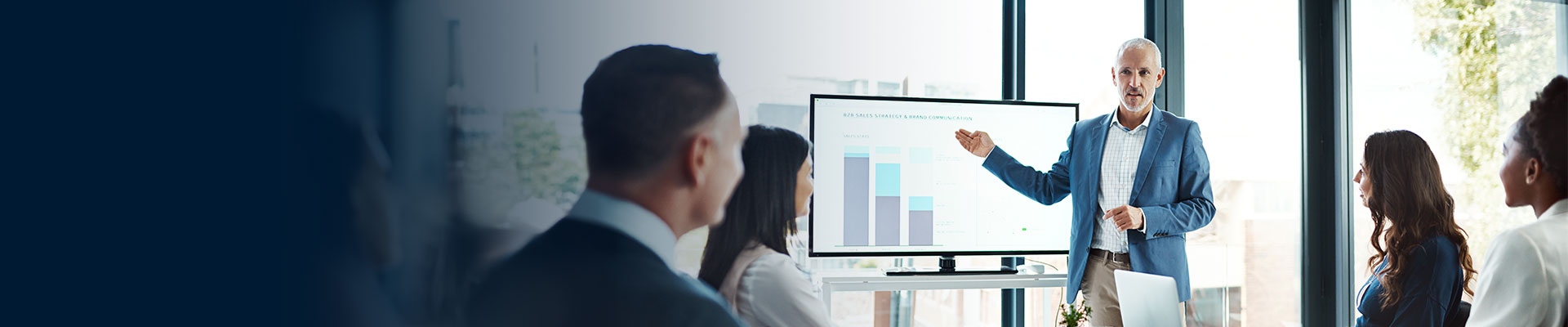 A professional setting with several people in business attire. One person is standing and presenting information on a large screen that displays bar graphs. The other individuals are seated and appear to be listening attentively to the presentation. The background includes large windows with natural light coming through.