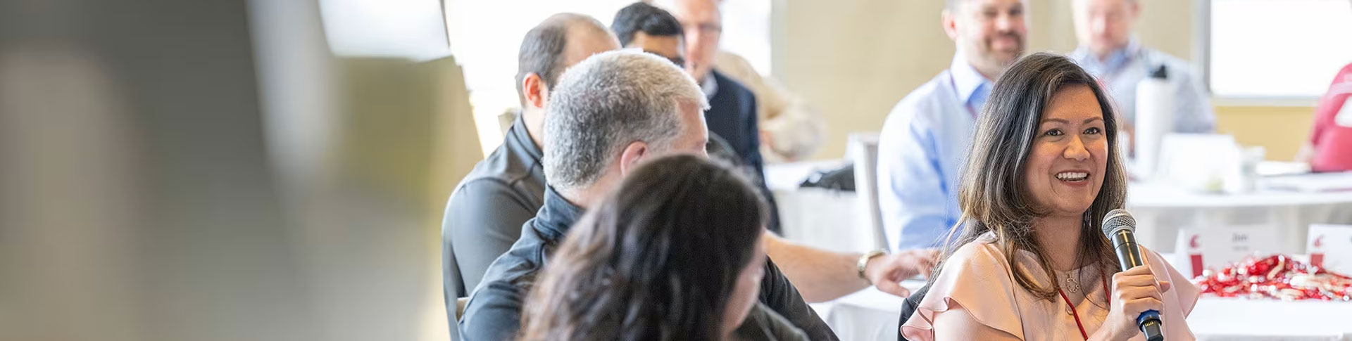 A group of people seated at tables during the EMBA Leadership Conference. One person in the foreground is holding a microphone and speaking, while others are listening attentively. The setting includes tables with white tablecloths and name tags, suggesting an organized event.