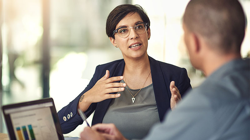 Two people engaged in a discussion. One person is gesturing with their hands while the other person is holding a pen and appears to be listening. There is a laptop on the table displaying a bar chart or graph.