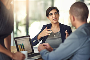 Two people engaged in a discussion. One person is gesturing with their hands while the other person is holding a pen and appears to be listening. There is a laptop on the table displaying a bar chart or graph.