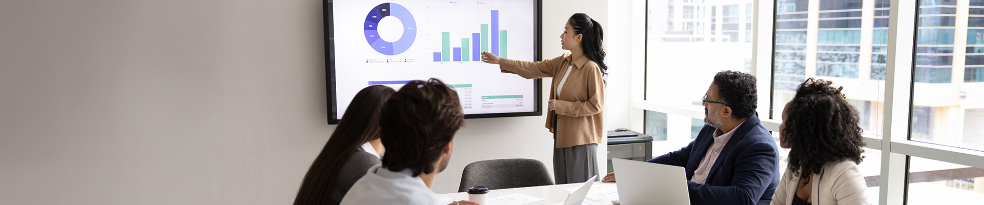 A business meeting with six individuals seated around a conference table. One person is standing and pointing at a large screen displaying various charts and graphs. The screen includes a pie chart, bar graph, and other data visualizations. There are laptops, documents, coffee cups, and other office supplies on the table. The room has large windows allowing natural light to enter.