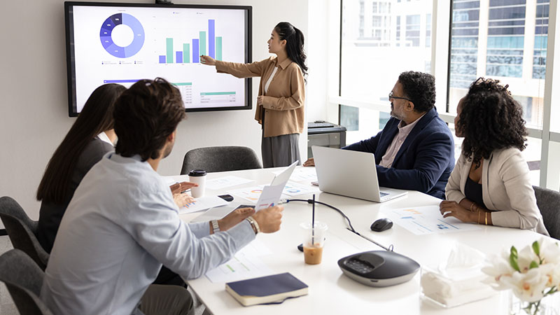 A business meeting with six individuals seated around a conference table. One person is standing and pointing at a large screen displaying various charts and graphs. The screen includes a pie chart, bar graph, and other data visualizations. There are laptops, documents, coffee cups, and other office supplies on the table. The room has large windows allowing natural light to enter.