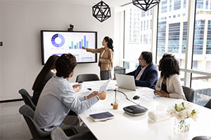 A business meeting with six individuals seated around a conference table. One person is standing and pointing at a large screen displaying various charts and graphs. The screen includes a pie chart, bar graph, and other data visualizations. There are laptops, documents, coffee cups, and other office supplies on the table. The room has large windows allowing natural light to enter.