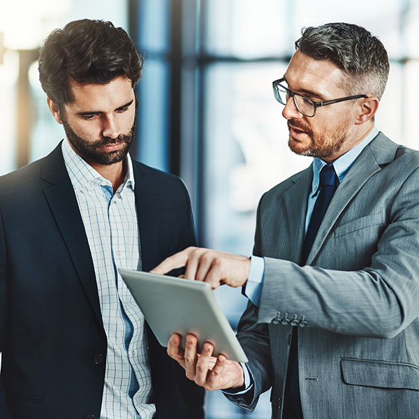 Two individuals in business attire, one holding a tablet and pointing at it while the other looks on. The background appears to be an office setting with large windows.