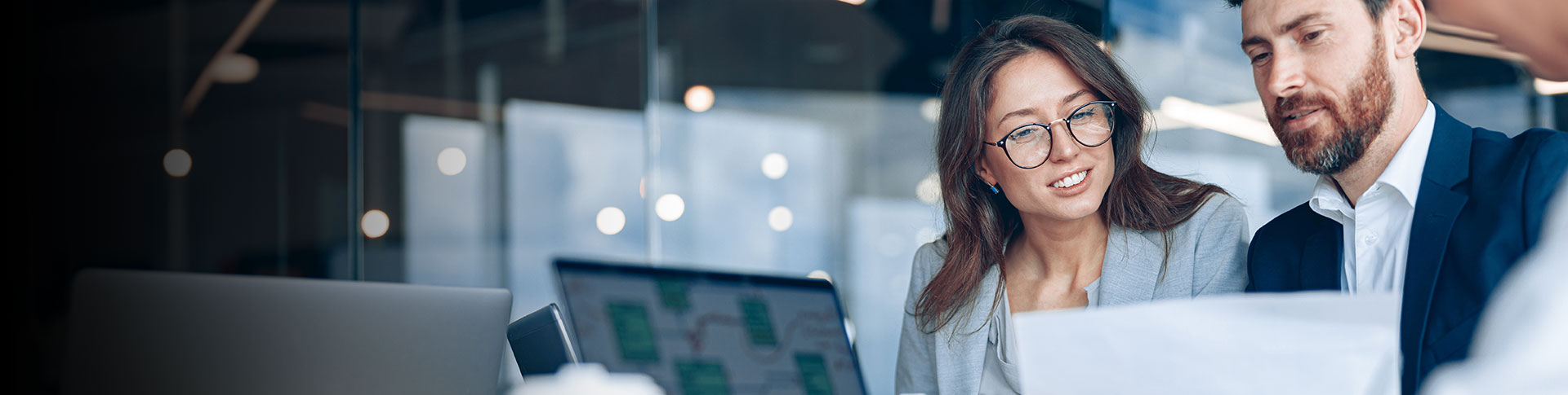 Two individuals sitting at a desk in an office setting, looking at some documents or papers. There is a laptop open on the desk in front of them. The background includes glass walls and reflections of lights, suggesting a modern office environment.