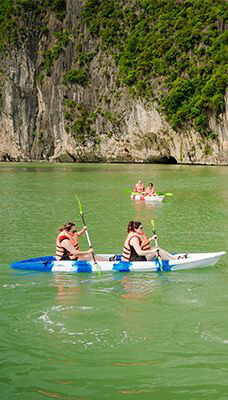 Two kayaks on the green waters of Ha Long Bay, Vietnam. The foreground features a white kayak with two people paddling, both wearing life jackets. In the background, another kayak with three people is visible. The backdrop consists of steep limestone cliffs covered in lush greenery.