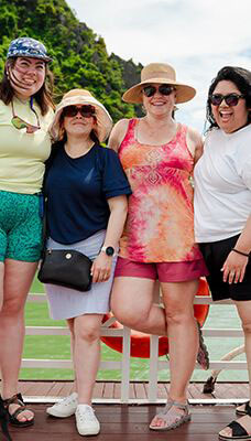 Four people are standing on a boat with a scenic background of Ha Long Bay in Vietnam. They are dressed in casual summer clothing. The background features lush green hills and calm water.