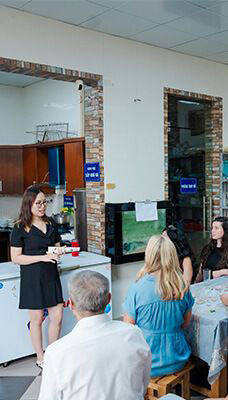 A group of people gathered in what appears to be a kitchen or dining area at Hope Box Vietnam, a social enterprise supporting women who have experienced gender-based violence. A woman is standing and speaking to the group, holding some papers or cards. The other individuals are seated around a table, listening to her. The background includes kitchen cabinets, appliances, and an aquarium. There are also blue signs with white text on the walls.