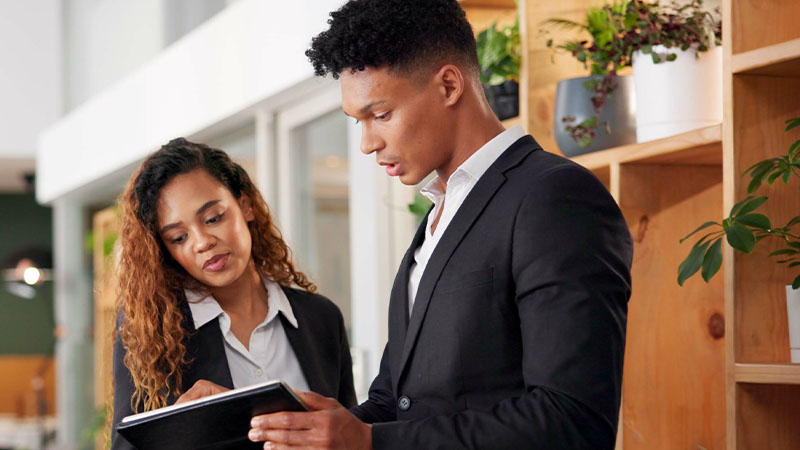 Two individuals dressed in formal business attire, standing in what appears to be a professional setting. One individual is holding and looking at a tablet or notebook, while the other is looking at the same device. There are shelves with potted plants and other items in the background.