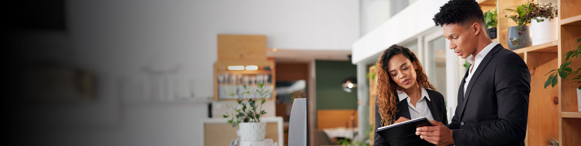 Two individuals dressed in formal business attire, standing in what appears to be a professional setting. One individual is holding and looking at a tablet or notebook, while the other is looking at the same device. There are shelves with potted plants and other items in the background.