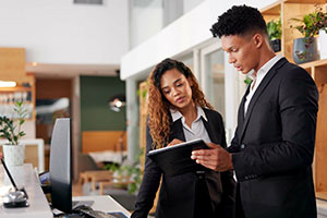 Two individuals dressed in formal business attire, standing in what appears to be a professional setting. One individual is holding and looking at a tablet or notebook, while the other is looking at the same device. There are shelves with potted plants and other items in the background.