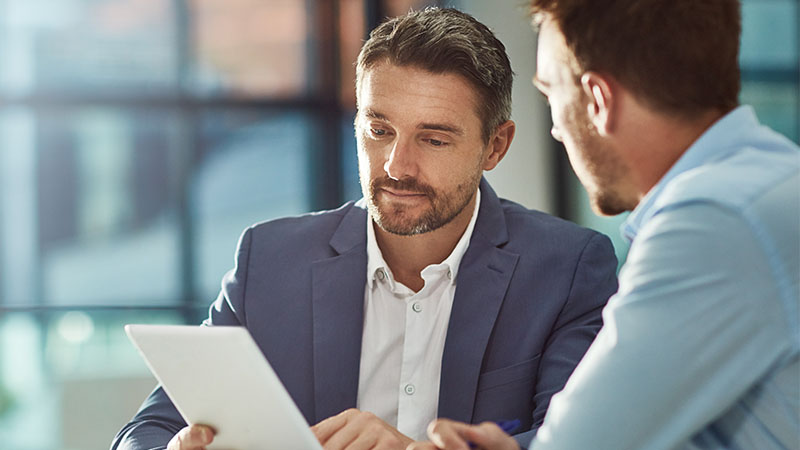 Two individuals sitting at a table, engaged in a discussion. One person is holding a tablet or document. Both individuals' faces are blurred out. The background appears to be an office setting with large windows and natural light.
