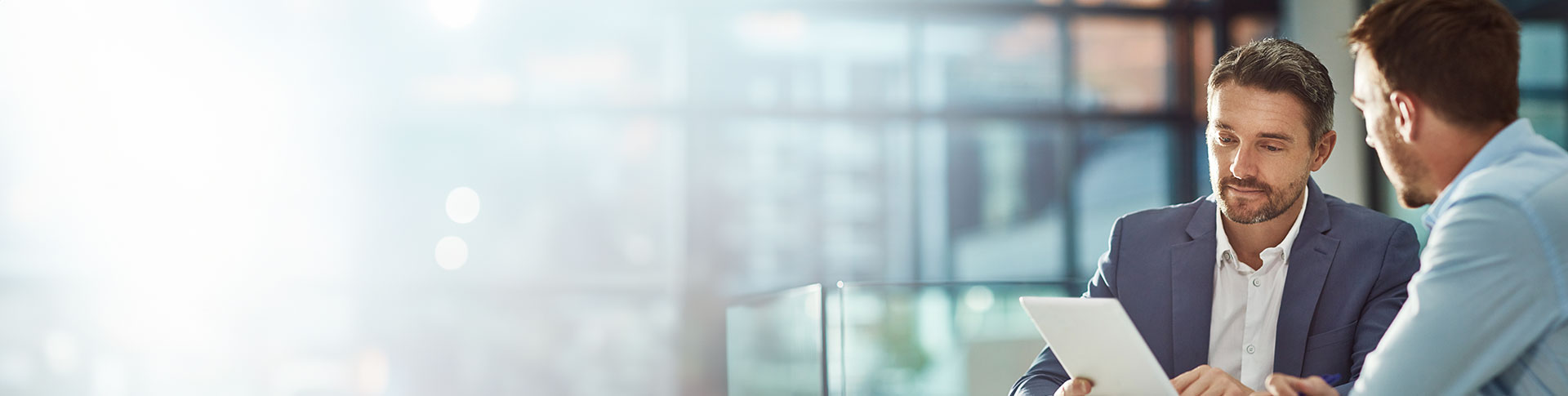 Two individuals sitting at a table, engaged in a discussion. One person is holding a tablet or document. Both individuals' faces are blurred out. The background appears to be an office setting with large windows and natural light.