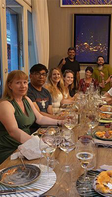 A group of people sitting around a dining table, enjoying a meal together. The table is set with plates of food, wine glasses, and bottles. There is a large window on the left side and a framed picture on the wall in the background. The location is indoors in Italy.