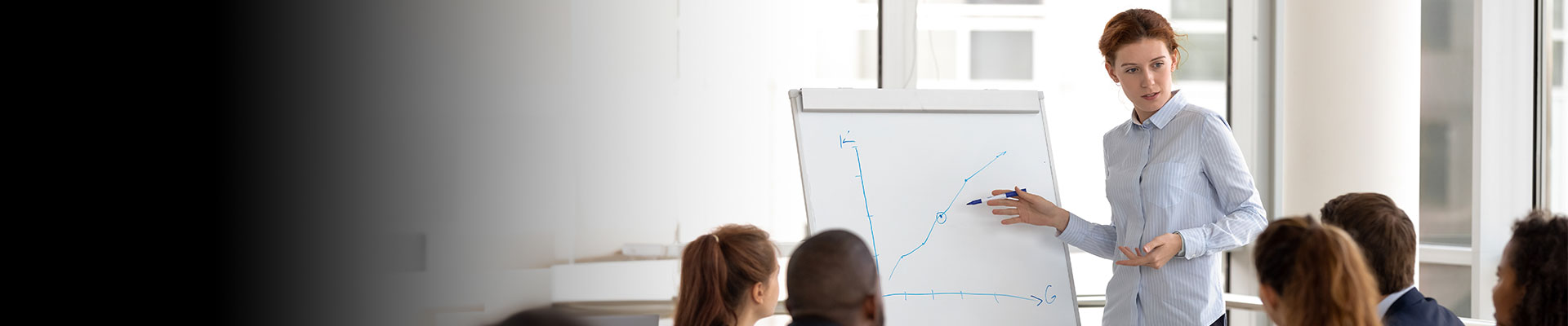 A person standing in front of a whiteboard, giving a presentation to an audience. The whiteboard has some graphs drawn on it with blue marker. There are several people seated and facing the presenter, indicating that this is likely a professional or educational setting.