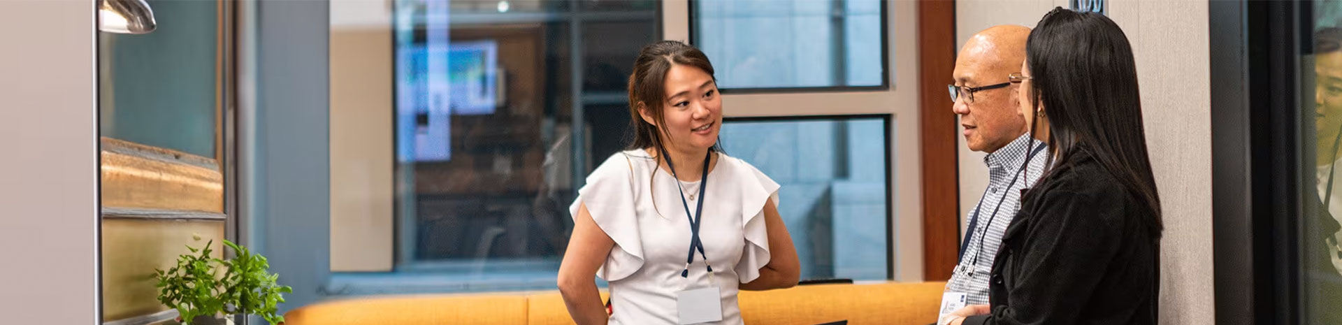 Three people standing and conversing in what appears to be an office or a professional setting. There is a plant on the left side, and the background includes windows with reflections of other parts of the office.
