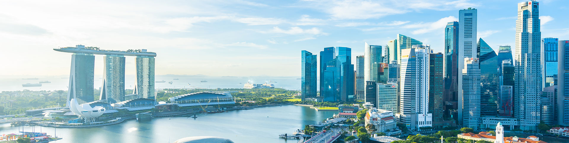 A panoramic view of a modern cityscape with numerous high-rise buildings and skyscrapers. There is a large body of water in the foreground, and the skyline includes notable architectural structures such as the Marina Bay Sands hotel complex on the left side. The sky is clear with some clouds, suggesting a bright and sunny day.