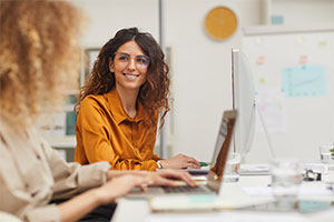 Two people working at desks with computers. The person in the foreground is typing on a laptop, while the person in the background is looking at a desktop monitor. The background includes some office supplies and a whiteboard with notes and charts.