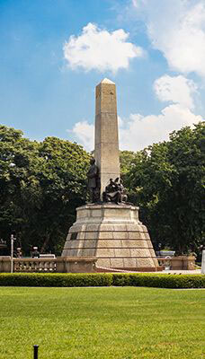 The Rizal Monument located in Rizal Park, Manila, Philippines. It features a bronze sculpture of Dr. José Rizal, a Filipino nationalist and revolutionary, standing on a pedestal with an obelisk behind him. The structure is surrounded by a well-maintained garden and trees in the background.
