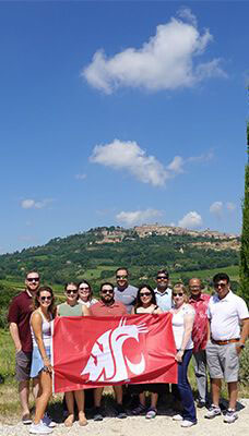 A group of people standing outdoors holding a red flag with a white Washington State University cougar head logo. The photo was taken at Salcheto Winery. The background features a scenic landscape with green hills and a small town or village on top of one of the hills under a blue sky with some clouds.