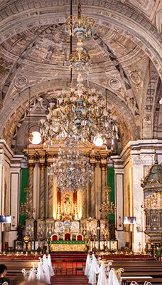 The interior of San Agustin Church, featuring an ornate altar adorned with gold and intricate decorations. Above the altar hangs a large, elaborate chandelier. The ceiling showcases detailed architectural designs and carvings. There are pews in the foreground, with some white draped decorations near the front rows.