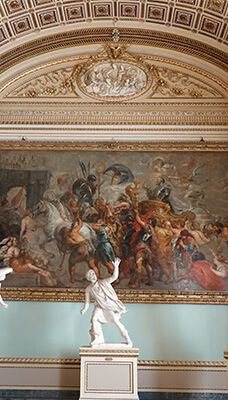 An interior view of the Uffizi Gallery in Italy. The focal point is a large, detailed painting depicting a historical or mythological scene with numerous figures, including warriors and horses, engaged in dynamic action. Below the painting are two white marble statues on pedestals. The ceiling above the painting is ornately decorated with intricate patterns and reliefs.