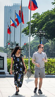 Two people walking on a paved path with several Philippine flags on flagpoles in the background. A tall building is visible in the distance, and trees and greenery surround the area. The person on the left is wearing a floral dress, while the person on the right is wearing a light-colored shirt and shorts.