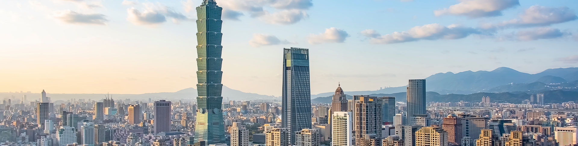 Taipei 101 rises above downtown Taipei, Taiwan, surrounded by modern high‑rises with mountains visible in the distance.