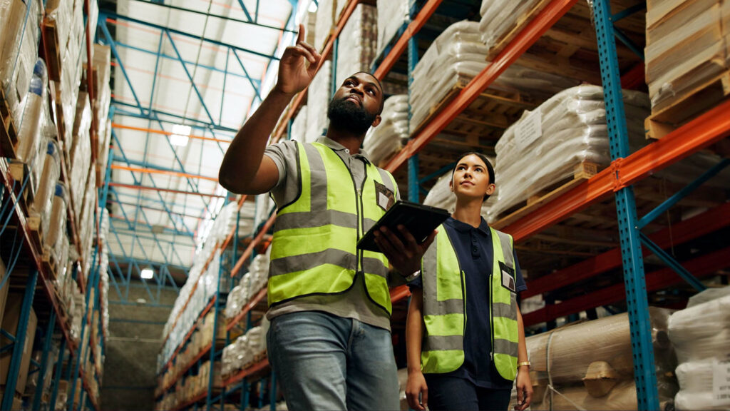 Two people in bright yellow vests walk through a warehouse, while one holds a tablet and points.