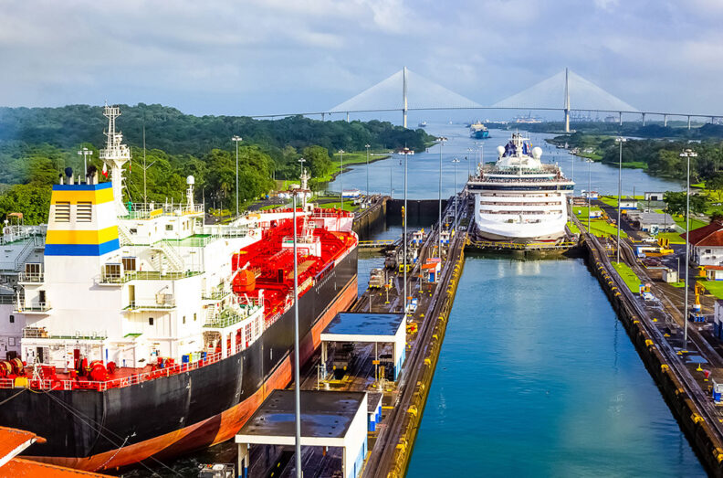 Two large ships passing through the Panama Canal, with surrounding greenery, infrastructure, and a bridge in the background.