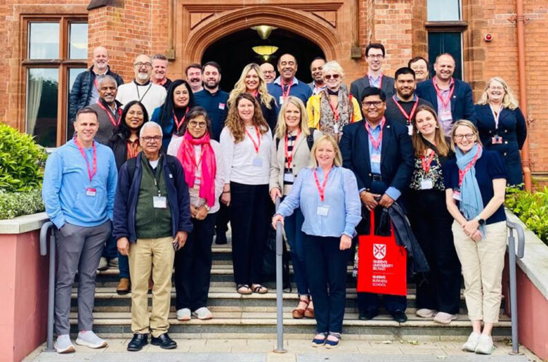 A large group of people stands on the steps of a historic brick building, posing together with name badges and red tote bags amid greenery.
