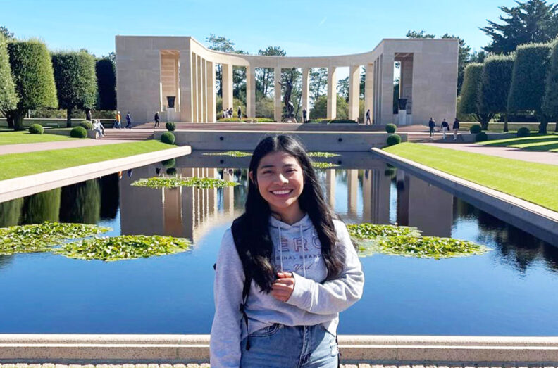 Hingry Mendez standing in front of the reflecting pool at the Normandy American Cemetery and Memorial, with the curved colonnade and landscaped grounds behind.